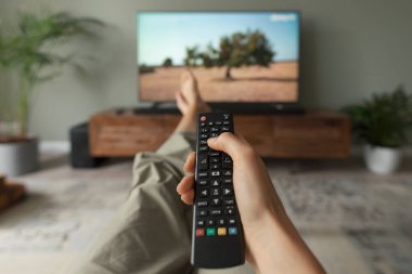 Evdeki kanepede televizyon izleyen ve uzaktan kumandayı tutan bir kadın.
