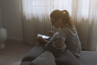 Genç ve çekici bir kadın oturma odasında dinleniyor, kanepede oturuyor ve dijital tabletine bağlanıyor.