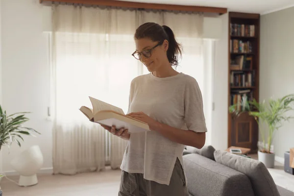 Happy woman standing in the living room and reading a book