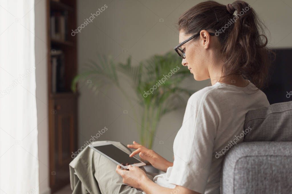 Young attractive woman relaxing at home in the living room, she is sitting on the couch and connecting online with her digital tablet