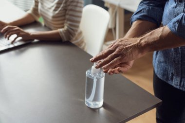 Ofiste birlikte çalışan iş adamları ve işyerindeki güvenlik önlemleri: bir erkek ellerine dezenfektan sürüyor