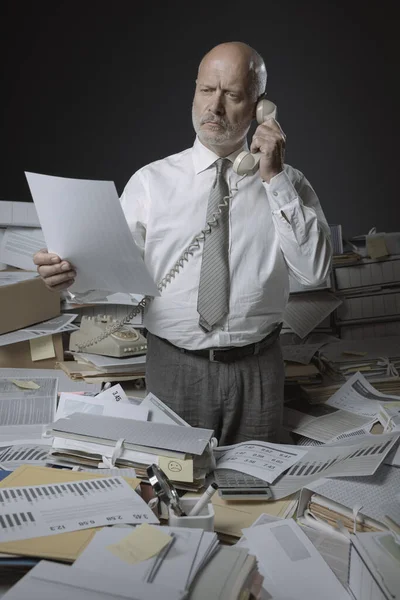 Confused stressed office worker answering phone calls, he is surrounded by lots of paperwork