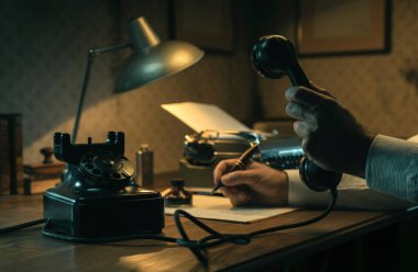 Ofisteki çalışma masasında çalışan kara film muhabiri elinde telefon ahizesini tutuyor ve notlar yazıyor.