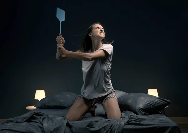 Angry woman killing mosquitoes in her bedroom, she is sitting on the bed and holding a fly swatter like a weapon