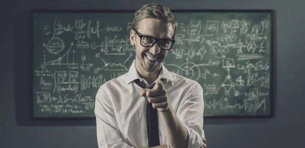 Confident smiling professor standing in front of a chalkboard with math formulas and pointing at camera