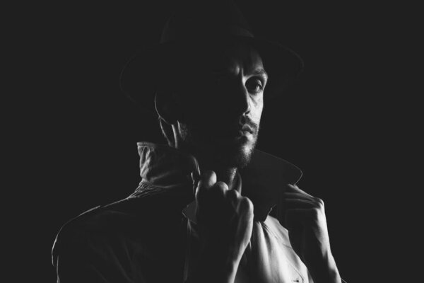 Noir film detective posing in the dark and staring at camera, thriller and crime concept