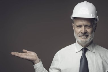 Gülümseyen orta yaşlı işadamı ve mühendis güvenlik kaskı takıyor ve kameraya bir şeyler sunuyor.