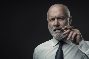 Kendinden emin üst düzey işletme yöneticisi puro içiyor ve kameraya bakıyor.