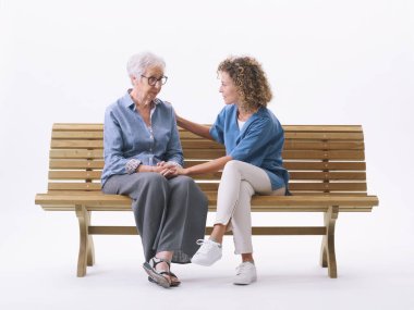 Profesyonel bir bakıcı, kafası karışmış yaşlı bir bayanı rahatlatıyor. Beyaz arka planda izole bir bankta oturuyorlar.