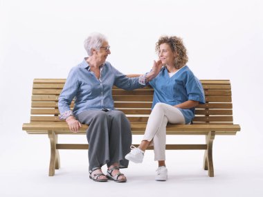 Bir hemşirenin yüzünü okşayan mutlu yaşlı bayan, beyaz arka planda soyutlanmış bir bankta oturuyorlar.