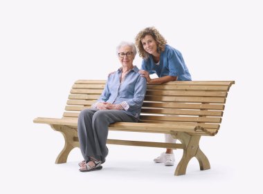 Mutlu yaşlı kadın ve bakıcı birlikte tahta bir bankta oturuyorlar, destekli yaşam konsepti, beyaz arka planda izole edilmiş.