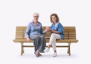 Dost canlısı hemşire yaşlı bir bayanla bankta oturuyor ve onu rahatlatıyor, elini tutuyor, beyaz arka planda tek başına.