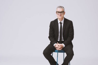 Taburede oturup kameraya bakan bir iş adamının portresi.