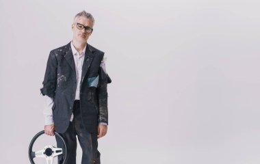 Orta yaşlı, yırtık elbiseli bir adamın komik bir portresi. Elinde araba direksiyonu, kırık camlar ve alnında bir çizik var. Tuhaf bir kaza, sakar bir başarısızlık ve beyaz arka planda komik bir felaketi temsil ediyor..