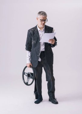 Sigorta sözleşmesini dikkatlice okurken elinde direksiyon tutan yırtık elbiseli bir adamın komik konsept görüntüsü. Araba kazası sembolü, sigorta terimleri, sigorta detayları ve dijital sigorta ve yol yardımının önemi.