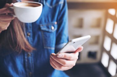 Bir kadının elinde tuttuğu, kahve içerken akıllı bir telefona dokunduğu yakın plan görüntüsü.