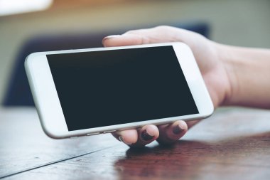 Beyaz cep telefonunu elinde tutan ve gösteren bir kadının resmi. Ahşap masadan izlemek için siyah ekranı boş.