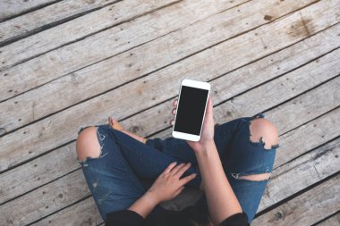 Üst görüntüde, eski ahşap zemin zemininde otururken elinde boş siyah ekranlı beyaz cep telefonu olan bir kadının görüntüsü var.