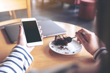 Kafede kek yerken elinde boş siyah masaüstü ekranı olan beyaz cep telefonu tutan bir kadının görüntüsü.