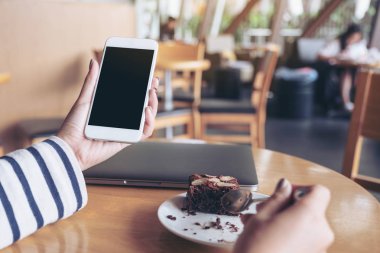Kafede kek yerken elinde boş siyah masaüstü ekranı olan beyaz cep telefonu tutan bir kadının görüntüsü.