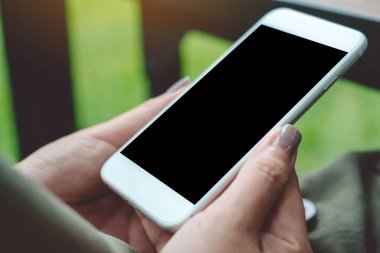 Beyaz akıllı telefonu olan, boş siyah masaüstü ekran ve bulanık yeşil arka planı olan bir kadının görüntüsü.