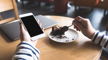 Kafede kek yerken elinde boş siyah masaüstü ekranı olan beyaz cep telefonu tutan bir kadının görüntüsü.