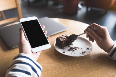 Kafede kek yerken elinde boş siyah masaüstü ekranı olan beyaz cep telefonu tutan bir kadının görüntüsü.