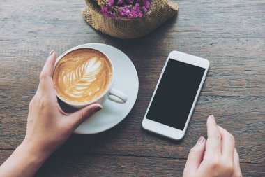 Mockup görüntü boş siyah perde ve sıcak latte kahve Cafe antika ahşap masa üzerinde tutan el ile beyaz cep telefonu