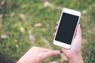 Mockup görüntüsünü tutan ve yeşil bahçe doğa arka planda boş siyah perde ile beyaz cep telefonu kullanarak bir kadın eli