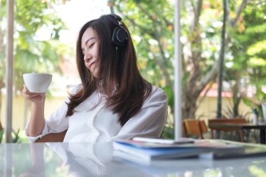 Yakın güzel bir Asya kadının gözleri ve kulaklık ile müzik dinlemek mutlu duygu ile içme kahve iken ve kafe yeşil doğa arka plan ile rahatlayın