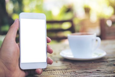 Mockup görüntüsünü holding beyaz cep telefonu ile boş ekran eller ve sıcak kahve Kupası tahta masada yeşil doğa arka plan bulanıklık 
