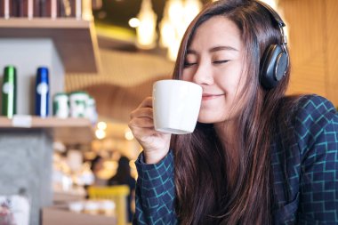 Güzel bir Asyalı kadın gözlerini kapatıp kulaklıkla müzik dinlerken modern kafede mutlu ve rahat bir şekilde kahve içiyor.