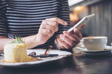 Ahşap masa restoranda pasta ve kahve Kupası ile akıllı telefon kullanan bir kadın 