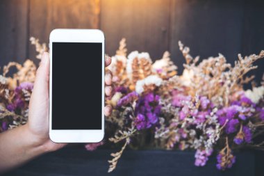 Mockup görüntü tutarak ve boş siyah perde ile renkli kuru çiçekler ve ahşap duvar arka plan beyaz cep telefonuyla kafede gösterilen El