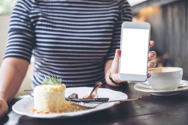 Bir kadın tutarak ve beyaz cep telefonu boş ekran, pasta ve kahve fincanı ile ahşap masa üzerinde modern kafede gösterilen mockup görüntü
