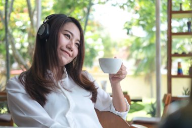 Güzel bir Asyalı kadın kulaklık ile müzik dinlemek süre mutlu duygu ile içme kahve ve kafe yeşil doğa arka plan ile rahatlayın