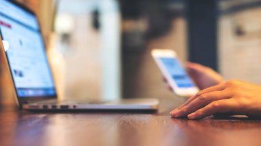 Ofiste ahşap tabloda dizüstü bilgisayar kullanırken cep telefonu tutan bir iş kadın