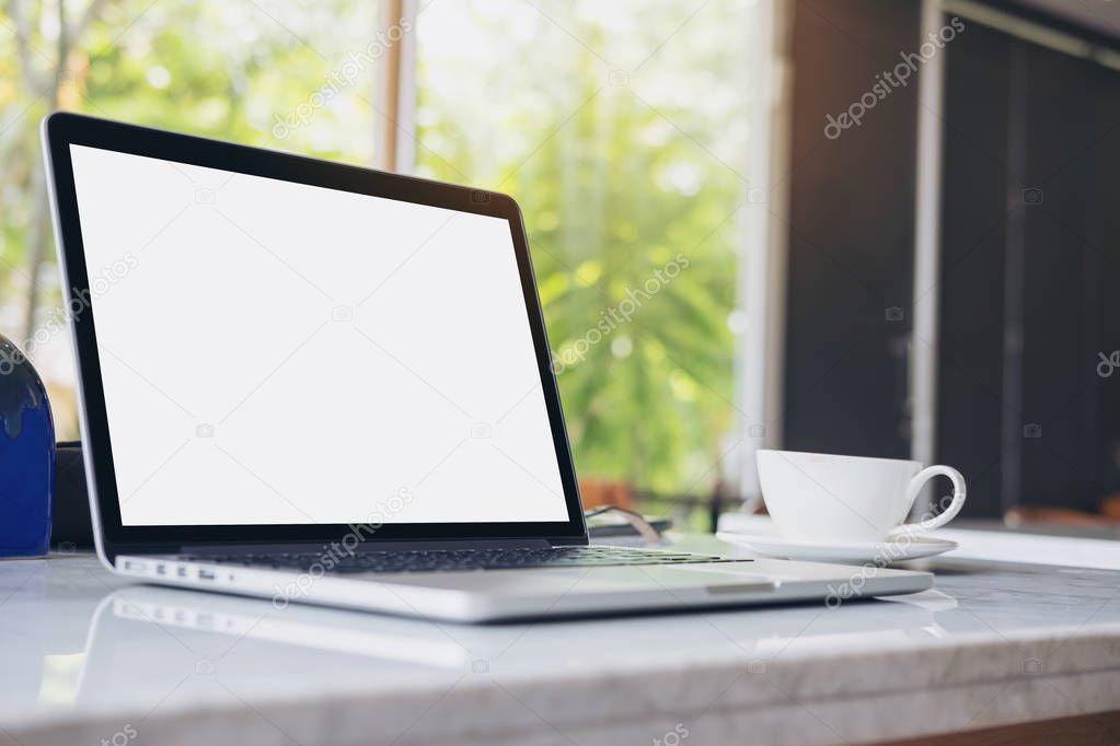 Mockup image of laptop with blank white screen and coffee cup on marble table in modern cafe with nature background