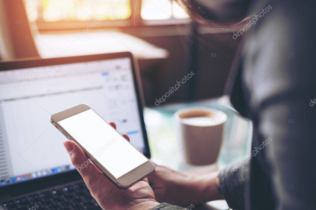 Mockup image of business woman holding mobile phone with blank white screen with latop on wooden table in cafe