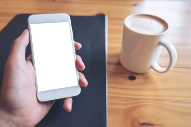 Kafede kahve fincanı ahşap tablo ile boş ekran ile beyaz cep telefonu tutan el mockup görüntü