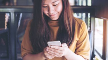 Kullanarak ve modern café'akıllı telefon bakarak gülen yüz güzel bir Asya kızla