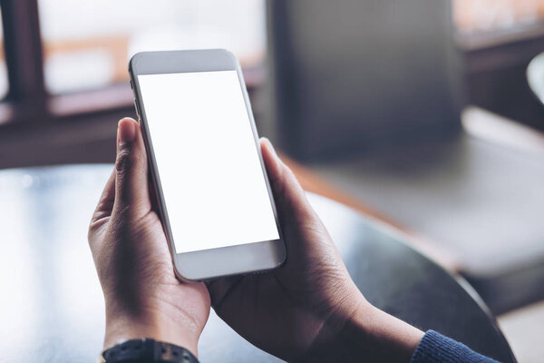 Mockup image of hands holding mobile phone with blank white screen in vintage cafe