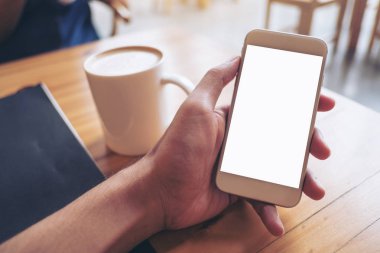 Kafede kahve fincanı ahşap tablo ile boş ekran ile beyaz cep telefonu tutan el mockup görüntü