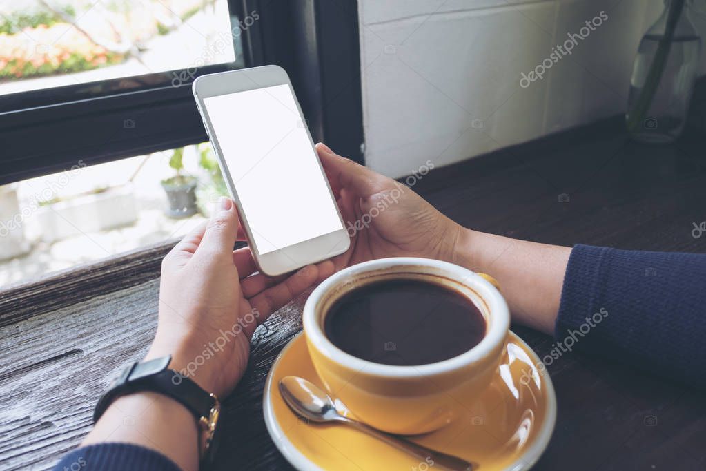 Mockup image of woman's hands holding white mobile phone with blank screen and yellow coffee cup on wooden table in cafe