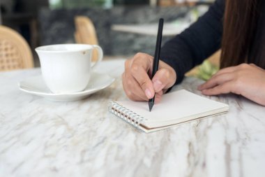 Kafede kahve fincanı ve tatlıyla beyaz boş bir deftere yazı yazan bir kadının ellerinin yakın plan resmi.
