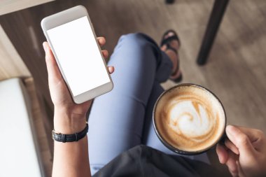 Kafede kahve içerken beyaz cep telefonu ve boş masaüstü ekranı olan bir kadının el görüntüsü.