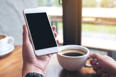 Kafede kahve içerken beyaz cep telefonunu siyah ekranla tutan ellerin görüntüsü.