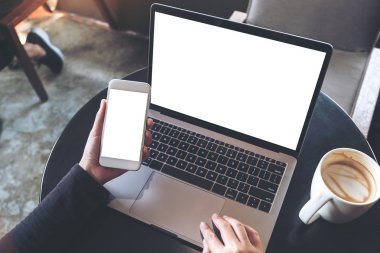 Masada boş beyaz masaüstü ekranı olan dizüstü bilgisayarı kullanırken boş cep telefonunu tutan ellerin en üstteki görüntüsü 