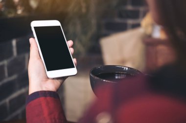Kafede kahve içerken beyaz cep telefonlu, siyah ekranlı bir kadının ellerinin görüntüsü.
