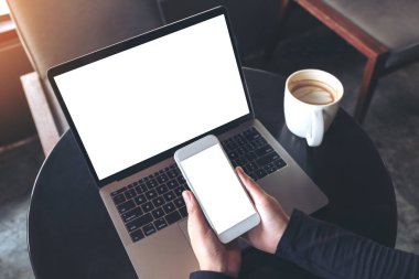 Masada boş beyaz masaüstü ekranı olan dizüstü bilgisayarı kullanırken boş cep telefonunu tutan ellerin en üstteki görüntüsü 
